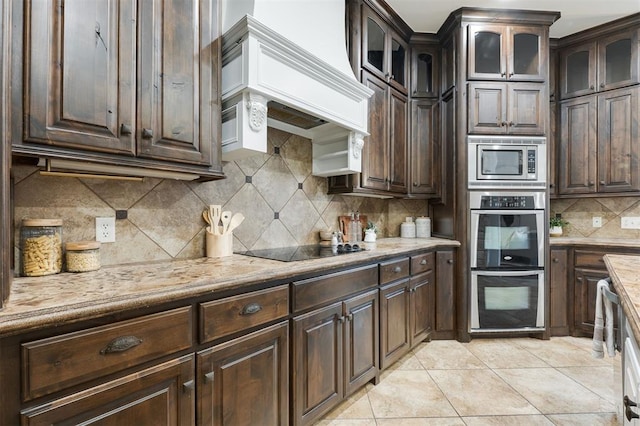 kitchen with light tile patterned floors, stainless steel appliances, dark brown cabinetry, custom range hood, and decorative backsplash