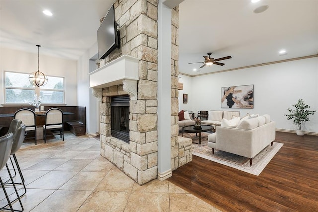 living room featuring crown molding, a stone fireplace, ceiling fan with notable chandelier, and light tile patterned floors