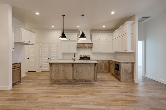 kitchen with white cabinetry, built in microwave, custom range hood, hanging light fixtures, and an island with sink