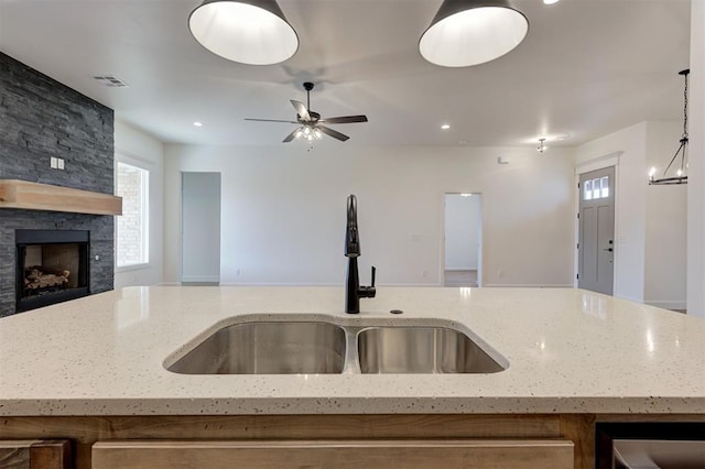 kitchen featuring sink, light stone counters, pendant lighting, a stone fireplace, and a kitchen island with sink