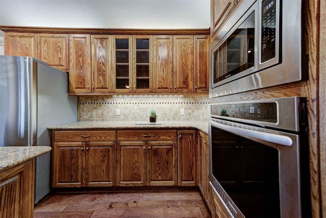 kitchen featuring brown cabinetry, glass insert cabinets, dark wood-type flooring, stainless steel appliances, and backsplash