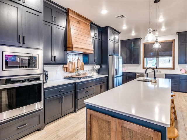 kitchen with premium range hood, appliances with stainless steel finishes, a breakfast bar, decorative light fixtures, and a center island with sink