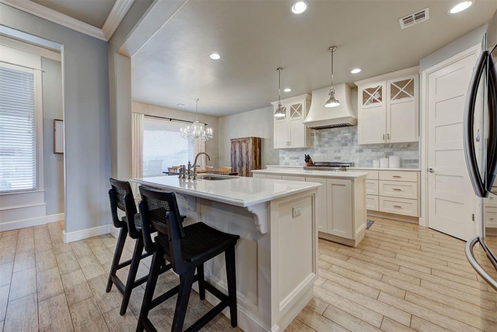 kitchen featuring a kitchen island with sink, white cabinetry, tasteful backsplash, decorative light fixtures, and custom exhaust hood