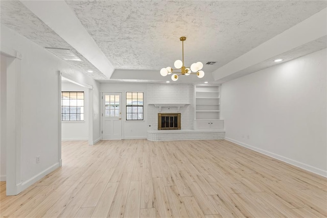 unfurnished living room with built in features, a notable chandelier, a brick fireplace, a raised ceiling, and light wood-type flooring