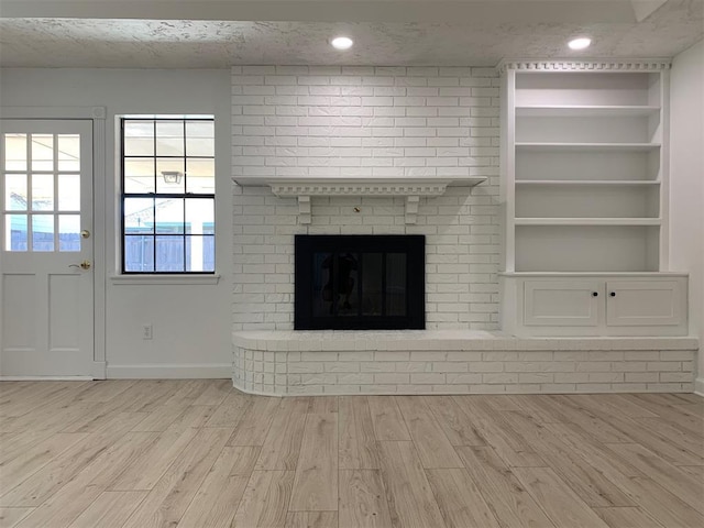 unfurnished living room with built in shelves, a textured ceiling, a brick fireplace, and light wood-type flooring