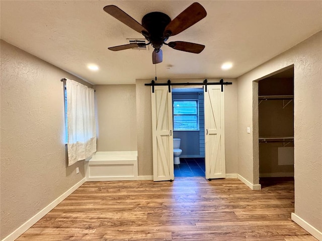 unfurnished bedroom featuring connected bathroom, a walk in closet, ceiling fan, a barn door, and light wood-type flooring