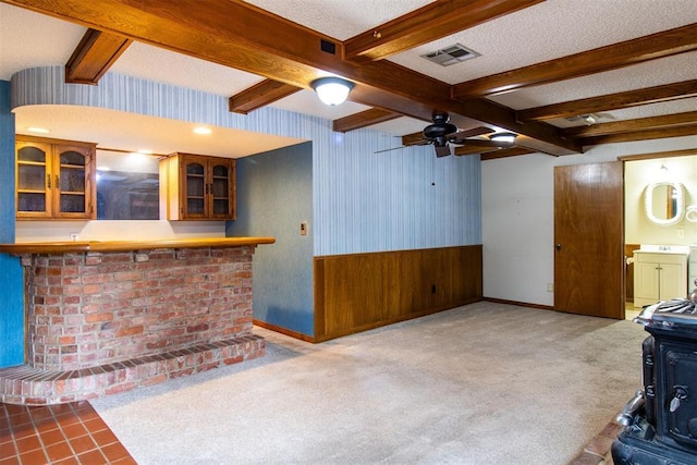 unfurnished living room featuring bar area, a textured ceiling, a wood stove, beamed ceiling, and light colored carpet