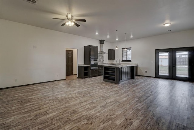 kitchen with hardwood / wood-style flooring, a center island with sink, decorative light fixtures, oven, and wall chimney exhaust hood