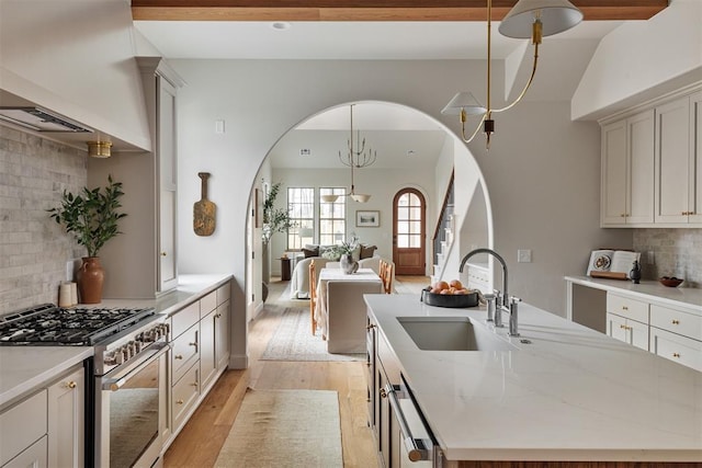 kitchen featuring an island with sink, sink, hanging light fixtures, light stone counters, and stainless steel gas range