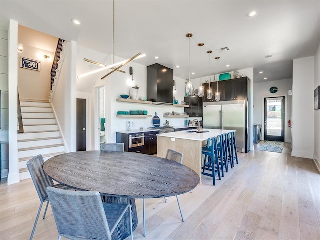 kitchen featuring built in refrigerator, light wood-type flooring, an island with sink, pendant lighting, and exhaust hood