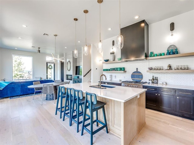 kitchen with a sink, light countertops, wall chimney exhaust hood, open shelves, and a center island with sink