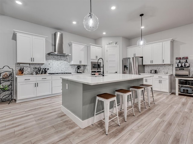 kitchen featuring decorative light fixtures, white cabinetry, a kitchen island with sink, stainless steel appliances, and wall chimney range hood