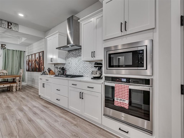 kitchen with wall chimney range hood, backsplash, stainless steel appliances, white cabinets, and light wood-type flooring