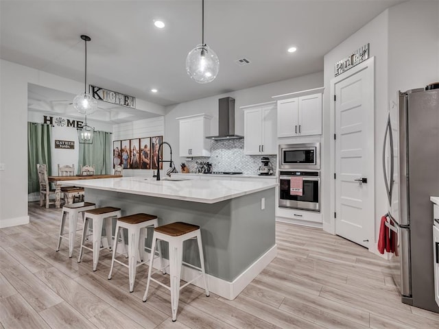 kitchen with appliances with stainless steel finishes, white cabinetry, sink, a center island with sink, and wall chimney exhaust hood