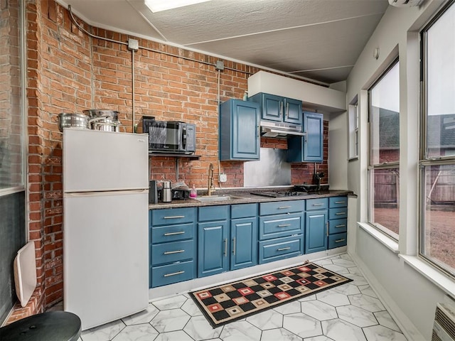 kitchen featuring brick wall, blue cabinets, stainless steel gas stovetop, sink, and white refrigerator