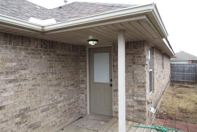 property entrance with brick siding, a shingled roof, and fence