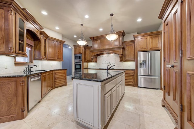 kitchen featuring decorative light fixtures, backsplash, dark stone counters, stainless steel appliances, and a center island with sink