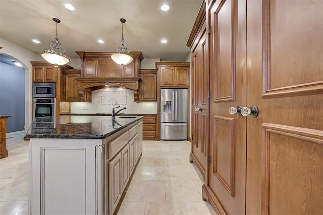 kitchen featuring appliances with stainless steel finishes, dark stone countertops, decorative backsplash, a center island with sink, and decorative light fixtures