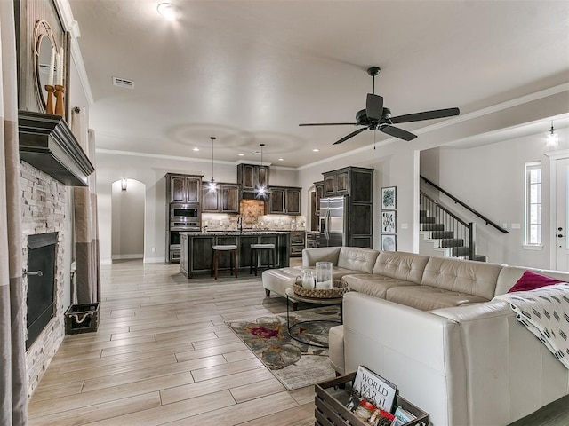 living room featuring sink, crown molding, ceiling fan, light hardwood / wood-style floors, and a stone fireplace