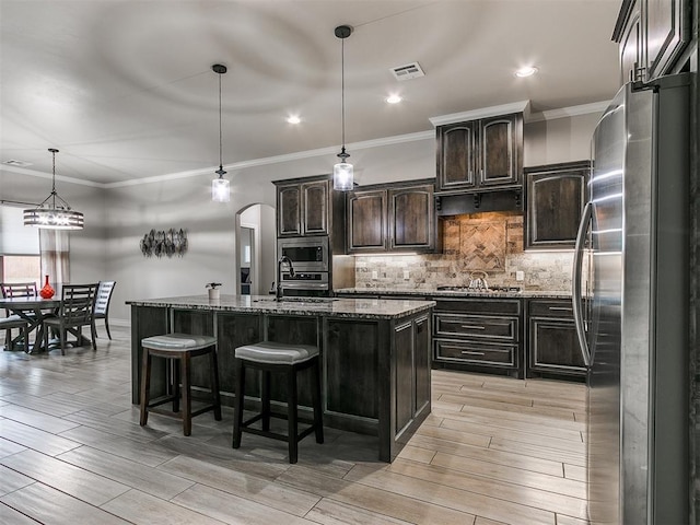 kitchen with dark brown cabinets, dark stone counters, an island with sink, pendant lighting, and stainless steel appliances