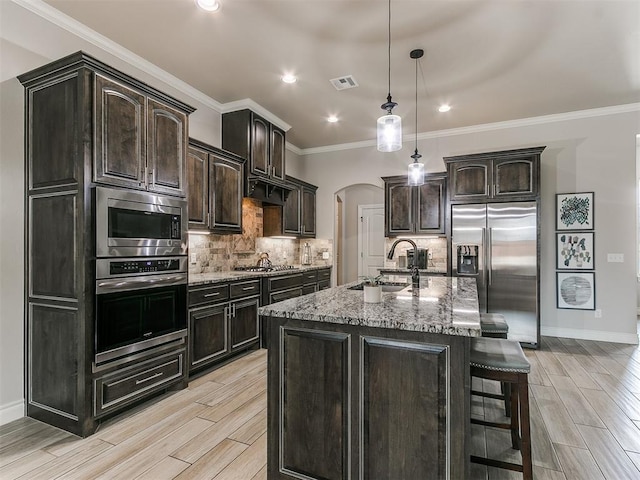 kitchen featuring appliances with stainless steel finishes, sink, dark brown cabinetry, light stone countertops, and a center island with sink