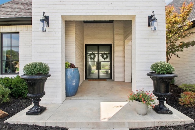 view of exterior entry featuring french doors, brick siding, and roof with shingles