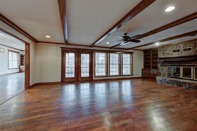 unfurnished living room featuring built in shelves, a stone fireplace, ornamental molding, beamed ceiling, and hardwood / wood-style floors