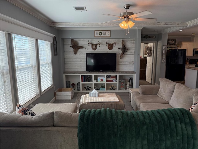 carpeted living room with ceiling fan, wooden walls, visible vents, and ornamental molding