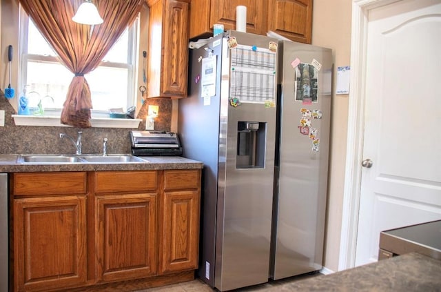 kitchen with brown cabinetry, a sink, decorative backsplash, dark countertops, and stainless steel fridge