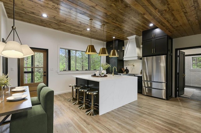 kitchen with stainless steel refrigerator, custom exhaust hood, hanging light fixtures, a kitchen island with sink, and wooden ceiling