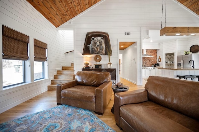 living room featuring light wood finished floors, wooden walls, vaulted ceiling, stairs, and wood ceiling