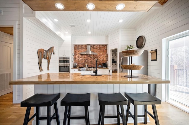 kitchen featuring wooden counters, wall chimney exhaust hood, light wood-style floors, stainless steel appliances, and wooden walls