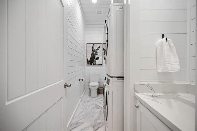 bathroom featuring estacked washer and dryer, marble look tiles, toilet, vanity, and wood walls