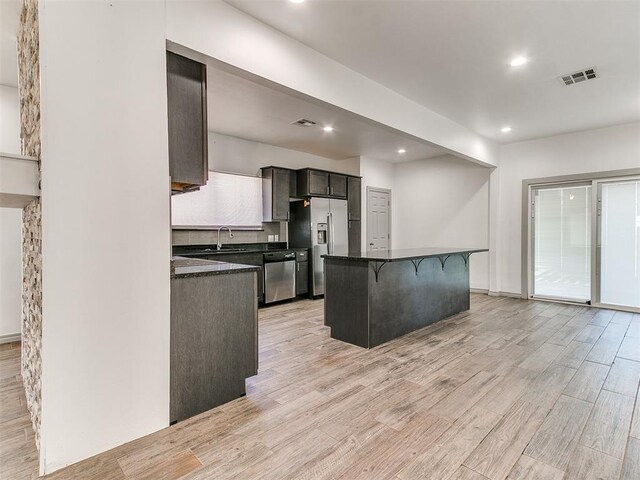kitchen with light wood finished floors, dark countertops, appliances with stainless steel finishes, a breakfast bar area, and a sink