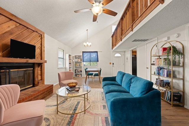 living room with ceiling fan with notable chandelier, high vaulted ceiling, a fireplace, hardwood / wood-style flooring, and a textured ceiling