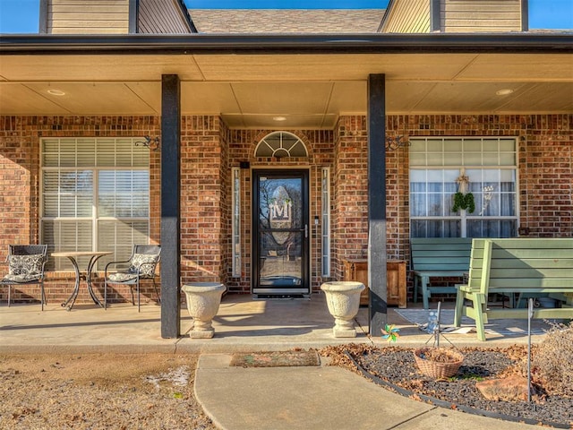 entrance to property featuring covered porch, brick siding, and roof with shingles