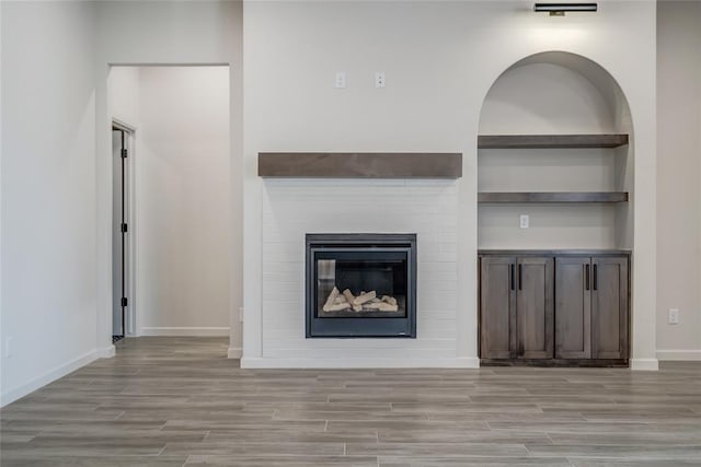 unfurnished living room featuring light wood finished floors, built in shelves, baseboards, and a glass covered fireplace