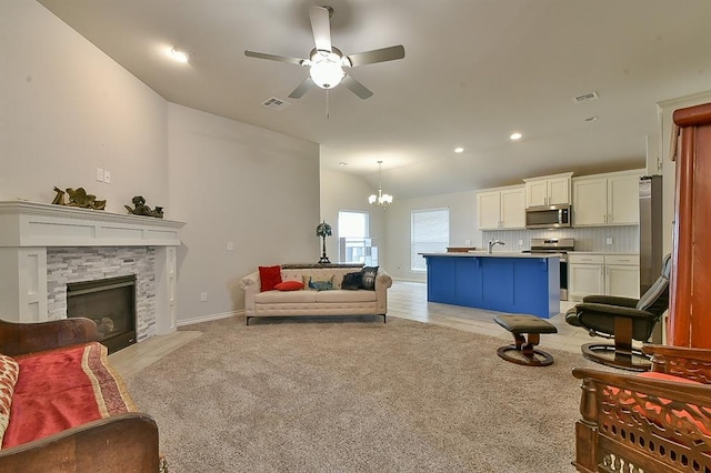 carpeted living room with sink, a stone fireplace, vaulted ceiling, and ceiling fan with notable chandelier