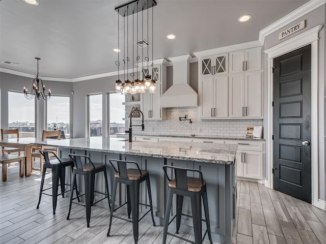 kitchen featuring white cabinetry, a kitchen breakfast bar, a large island with sink, hanging light fixtures, and custom range hood