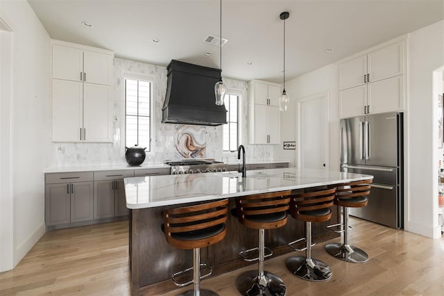 kitchen with stainless steel appliances, white cabinetry, an island with sink, premium range hood, and a kitchen breakfast bar