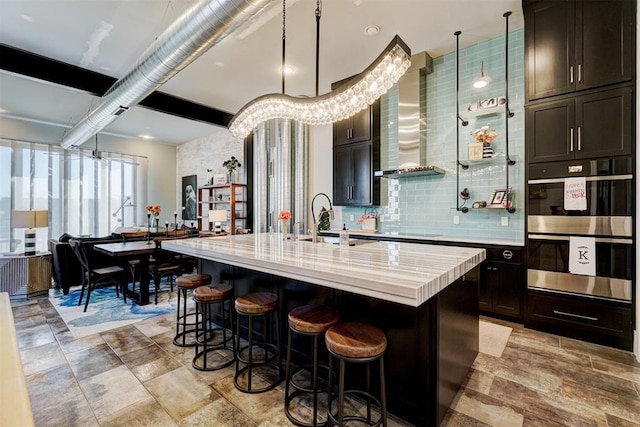 kitchen with decorative light fixtures, double oven, a center island with sink, and wall chimney range hood