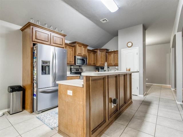 kitchen with light tile patterned floors, brown cabinetry, an island with sink, appliances with stainless steel finishes, and light countertops