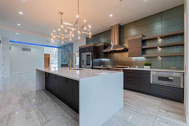 kitchen with sink, appliances with stainless steel finishes, wall chimney exhaust hood, decorative light fixtures, and a large island with sink