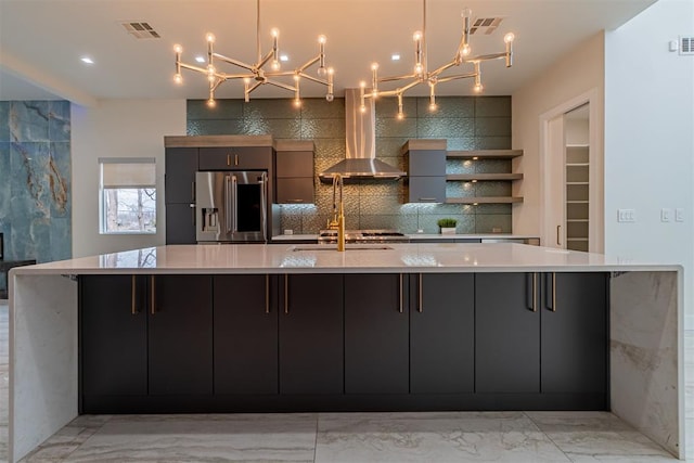 kitchen with pendant lighting, stainless steel fridge, wall chimney exhaust hood, decorative backsplash, and a large island with sink