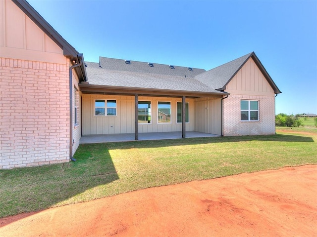 rear view of house featuring board and batten siding, a lawn, brick siding, a patio, and a shingled roof
