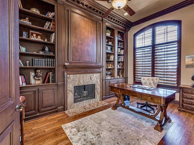 office area with built in shelves, wood finished floors, a high end fireplace, a ceiling fan, and ornamental molding