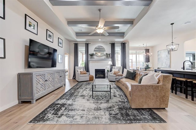 living area featuring light wood-style flooring, ceiling fan with notable chandelier, baseboards, a tray ceiling, and a glass covered fireplace