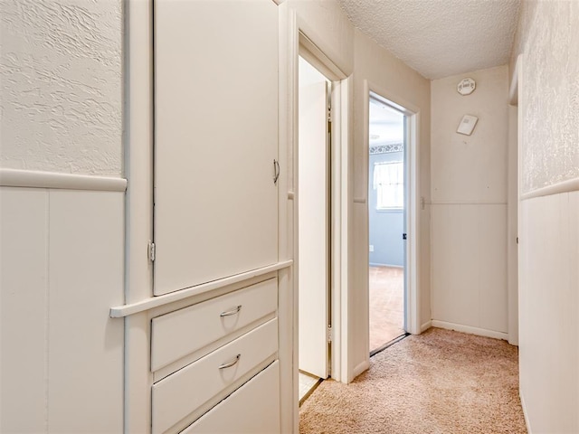 corridor with light carpet, a textured wall, a textured ceiling, and wainscoting