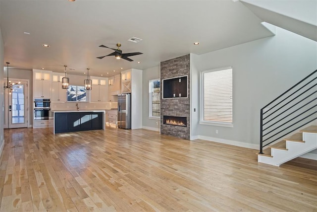 unfurnished living room with sink, a stone fireplace, light hardwood / wood-style floors, and ceiling fan with notable chandelier