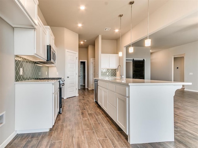 kitchen featuring appliances with stainless steel finishes, an island with sink, pendant lighting, a barn door, and white cabinetry